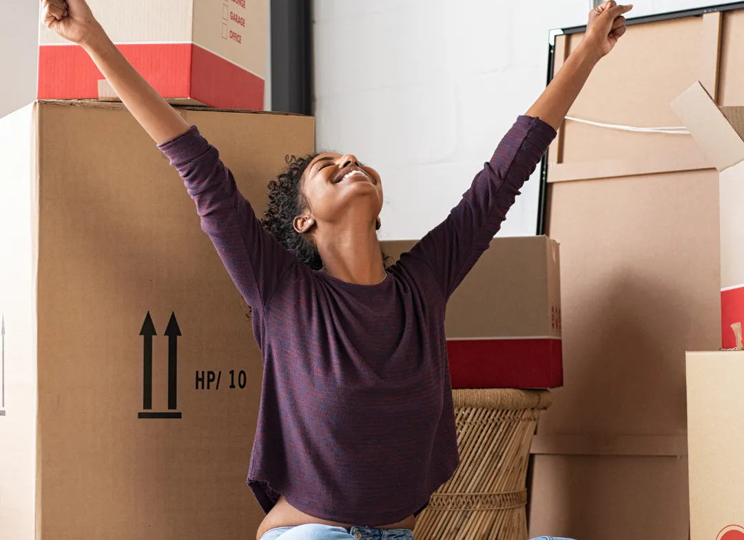 Mujer levanta los brazos de felicidad, con cajas de trasteo detrás. Invierte en vivienda en Colombia