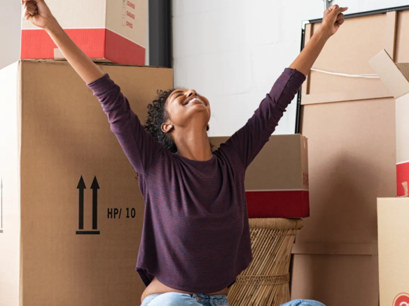 Mujer levanta los brazos de felicidad, con cajas de trasteo detrás. Invierte en vivienda en Colombia