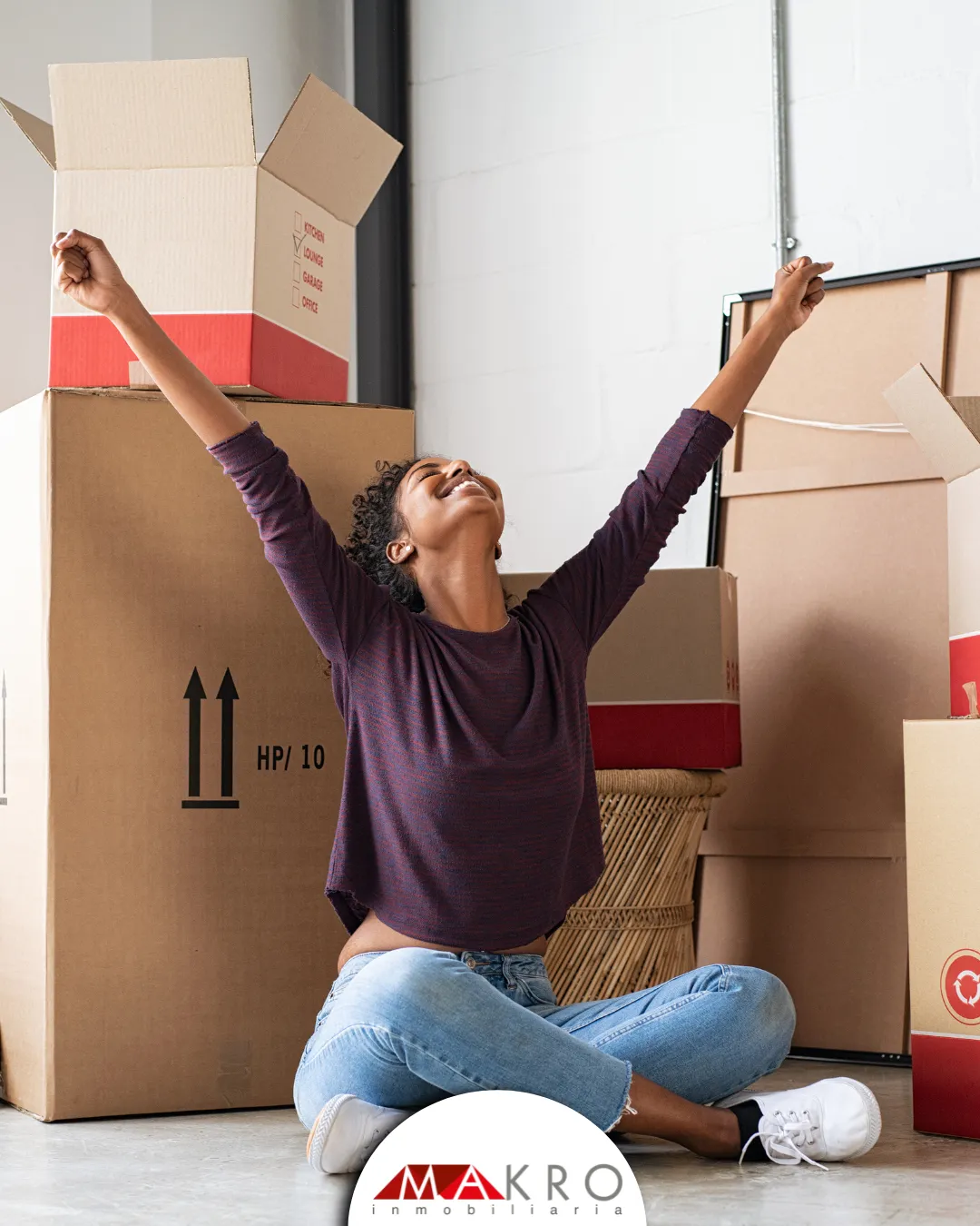 Mujer levanta los brazos de felicidad, con cajas de trasteo detrás. Invierte en vivienda en Colombia